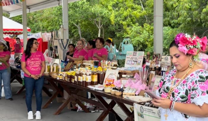 Vendedoras en stand de productos locales durante evento comunitario en Playa del Carmen con sombrillas y decoraciones rosa.