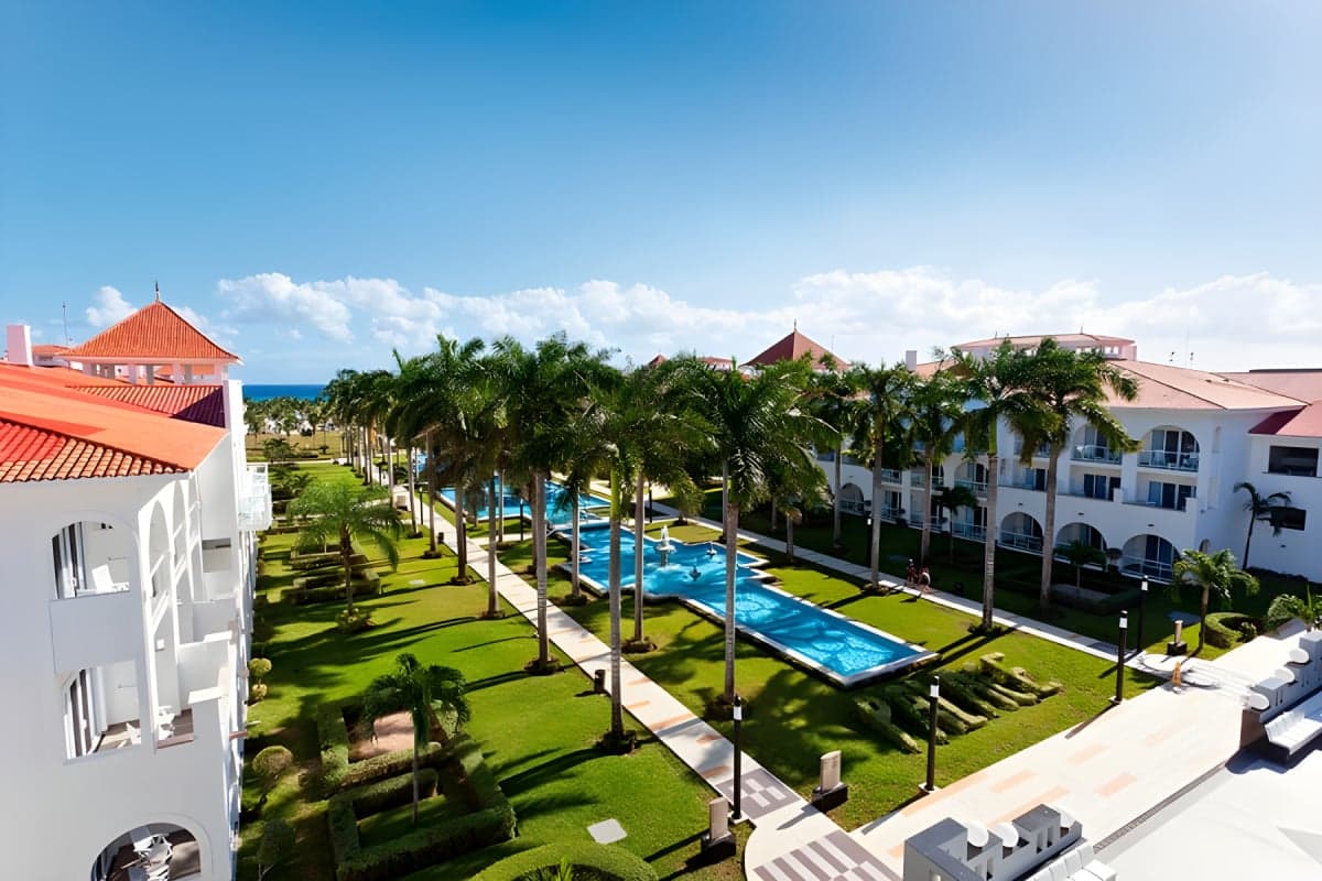 Vista aérea del resort Riu Palace México mostrando sus instalaciones renovadas con piscinas, áreas verdes, palmeras y edificios blancos frente al mar Caribe.