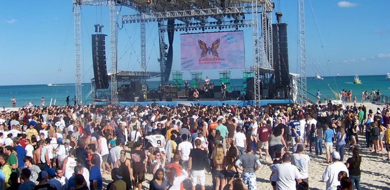 Multitud de personas en playa frente a escenario con pantalla gigante durante evento musical en la Riviera Maya.