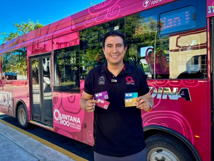 Persona posando frente a un autobús rosa de la ruta MOBI Quintana Roo, sosteniendo tarjetas de pago electrónico.