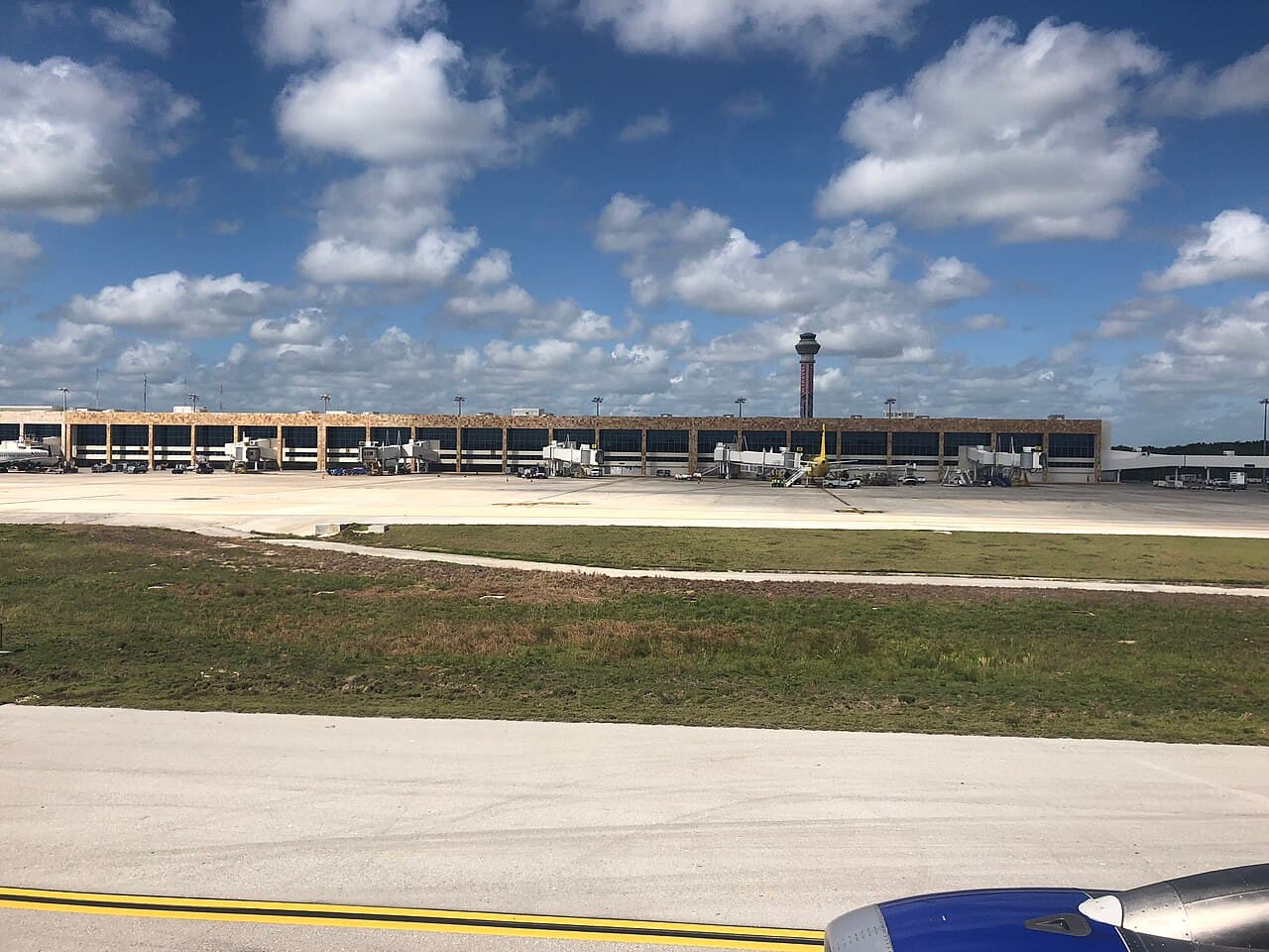 Vista desde la pista de una terminal de aeropuerto con varias posiciones de aeronaves, puentes de abordaje, vehículos de servicio y torre de control bajo un cielo parcialmente nublado.