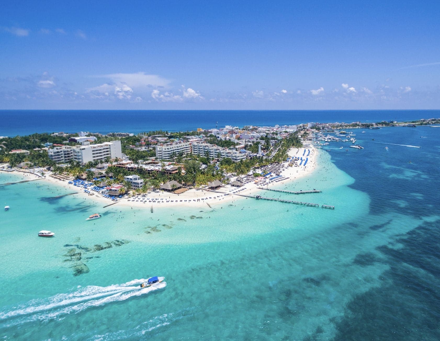 Vista aérea de una playa tropical con aguas turquesas cristalinas, arena blanca, resorts y embarcaciones, sin presencia visible de sargazo.