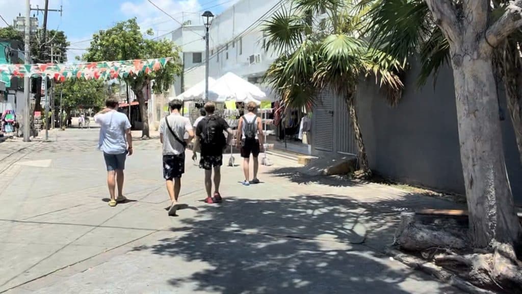 Varios turistas caminan por una calle del Centro de Cancún bajo un día soleado, con tiendas y una carpa blanca de evento visible de fondo.